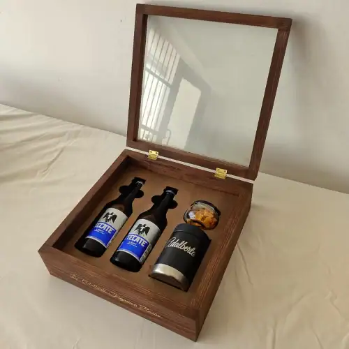 Un estuche de madera abierto, con la tapa de espejo que refleja una ventana. Dentro, se ven dos cervezas Tecate y un portacervezas con un frasco de botana.