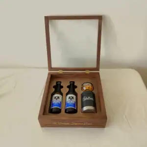 Un estuche de madera abierto con tapa de cristal, mostrando dos cervezas Tecate Light y un portacervezas negro con un frasco de botana, listo para regalar.