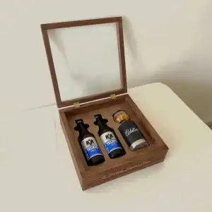 Un estuche de madera abierto, con la tapa de cristal levantada, mostrando dos cervezas Tecate y un portacervezas de metal con un frasco de botana en su interior.