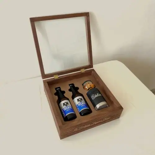Un estuche de madera abierto, con la tapa de cristal levantada, mostrando dos cervezas Tecate y un portacervezas de metal con un frasco de botana en su interior.