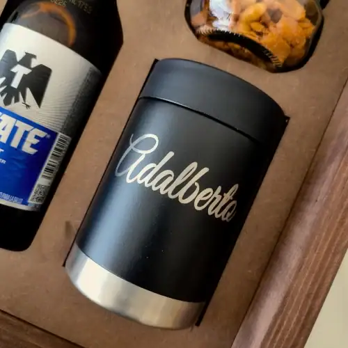 Primer plano de un portacervezas de metal negro con el nombre "Adalberto" grabado, dentro de un estuche de madera con una botella de cerveza.